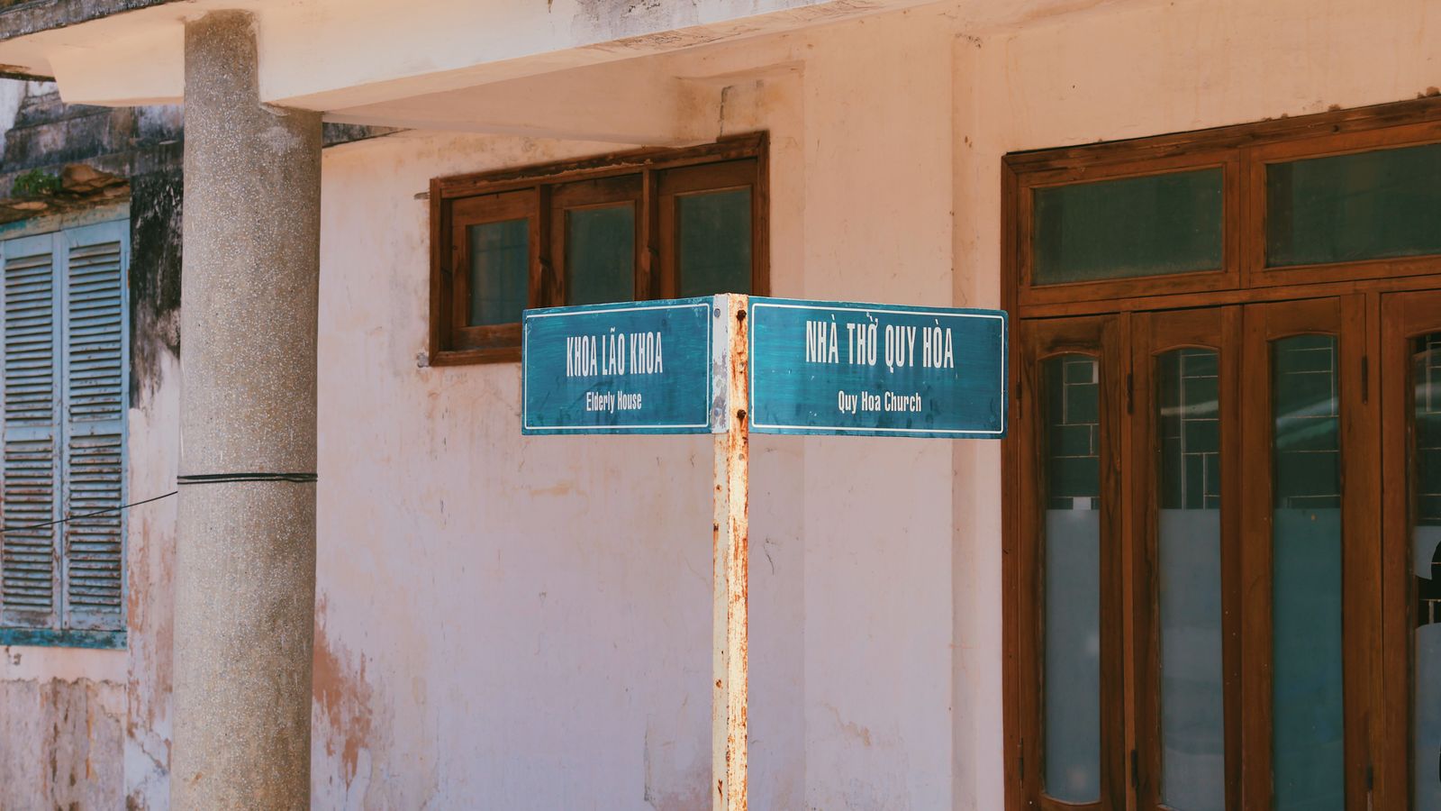 Two bilingual signposts on one rusting pole in a hospital compound — KHOA LAO KHOA, Elderly House, and NHA THO QUY HOA, Quy Hoa Church — against a peeling ochre wall