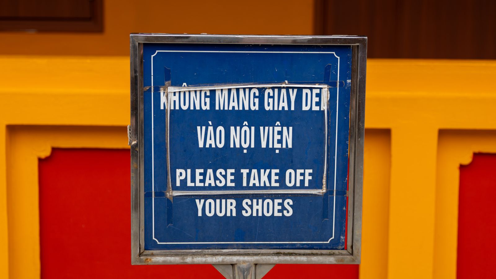 A blue enamel sign on a post in front of a red and yellow temple wall, reading KHONG MANG GIAY DEP VAO NOI VIEN above the English PLEASE TAKE OFF YOUR SHOES
