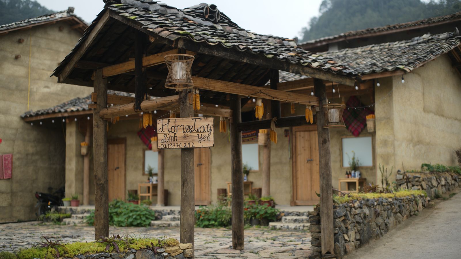A rammed-earth house behind a drystone wall, maize cobs and lanterns hung along its porch and a hand-painted board at the gate reading Homestay Binh Yen with a phone number