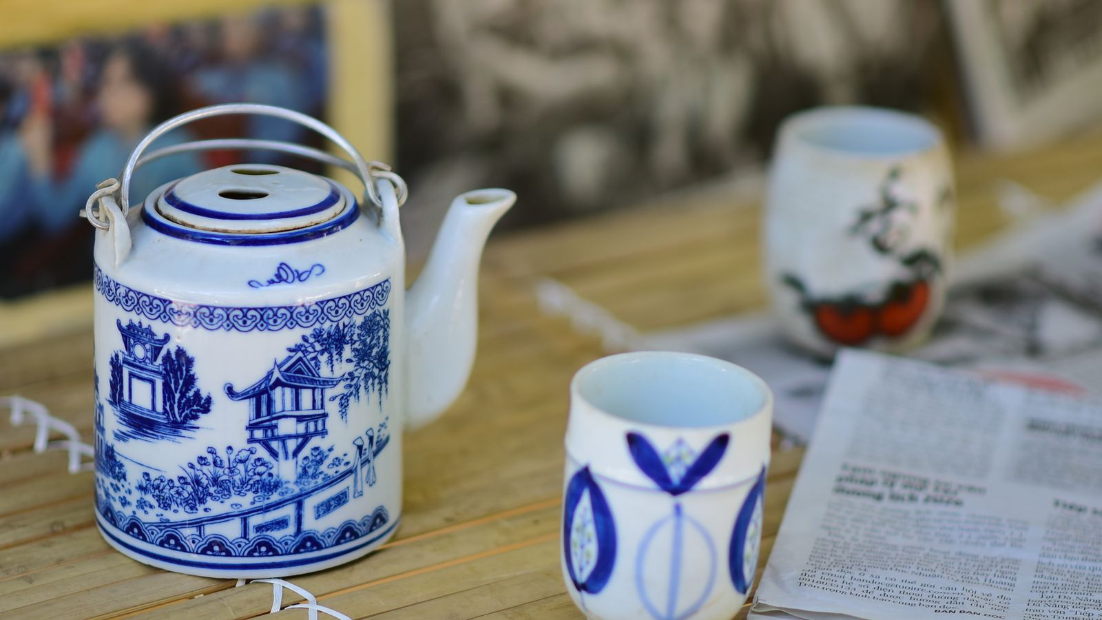 A blue and white Vietnamese teapot painted with the One Pillar Pagoda beside two small cups, a Vietnamese newspaper on the table