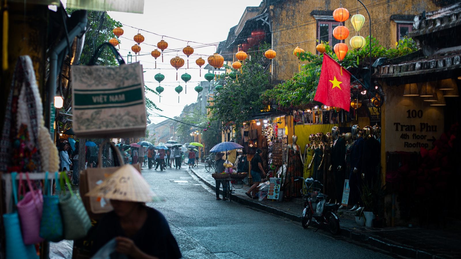 Lanterns strung across Tran Phu street in Hoi An in the rain, a Vietnamese flag on a yellow wall and a sign reading 100 Tran Phu