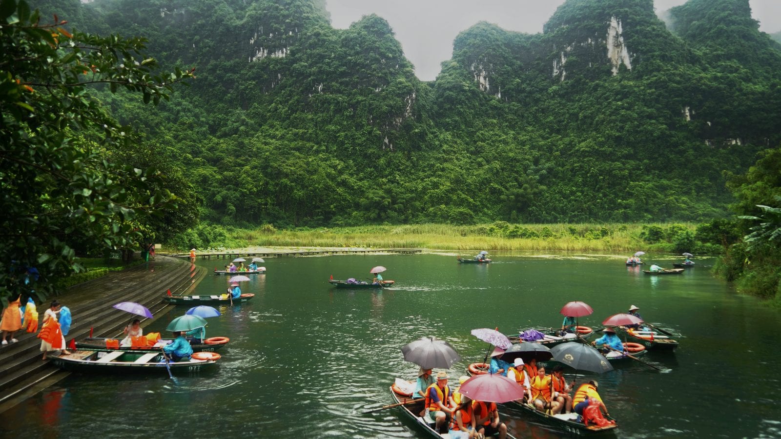 Numbered wooden rowing boats under umbrellas on a green karst river in the rain, rowers in conical hats and passengers in life jackets