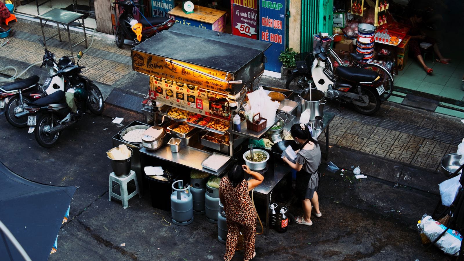 A street food cart seen from above, its glass case lit and a numbered menu board with prices in thousands stuck across the front, gas bottles and stock pots on the pavement beside it