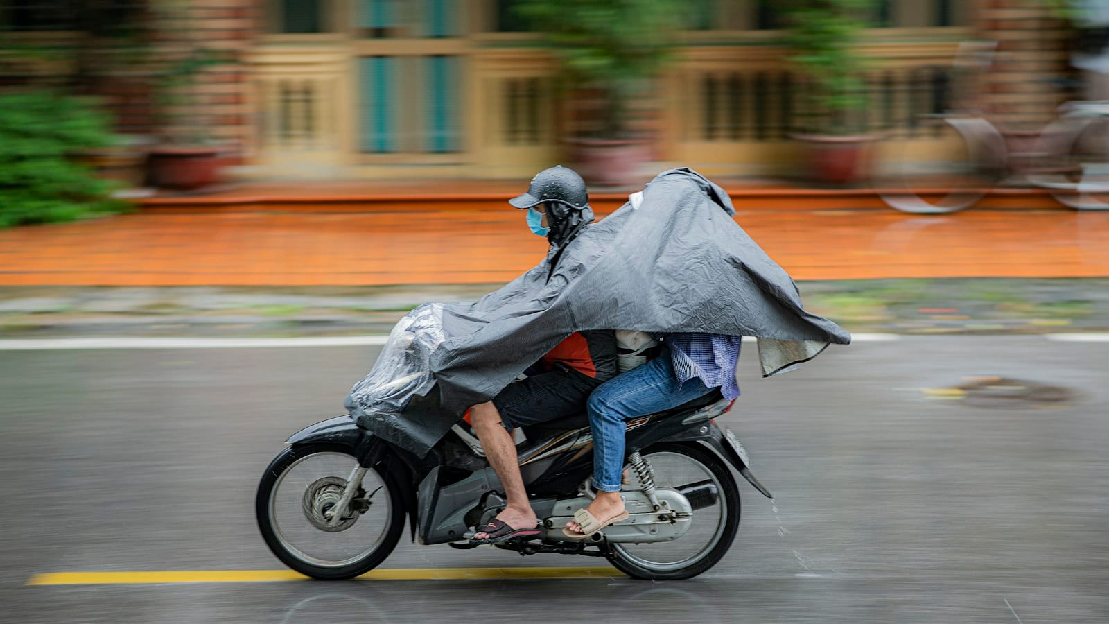 Two riders sharing one poncho stretched over both of them and the handlebars, panned against a yellow colonial wall in the wet