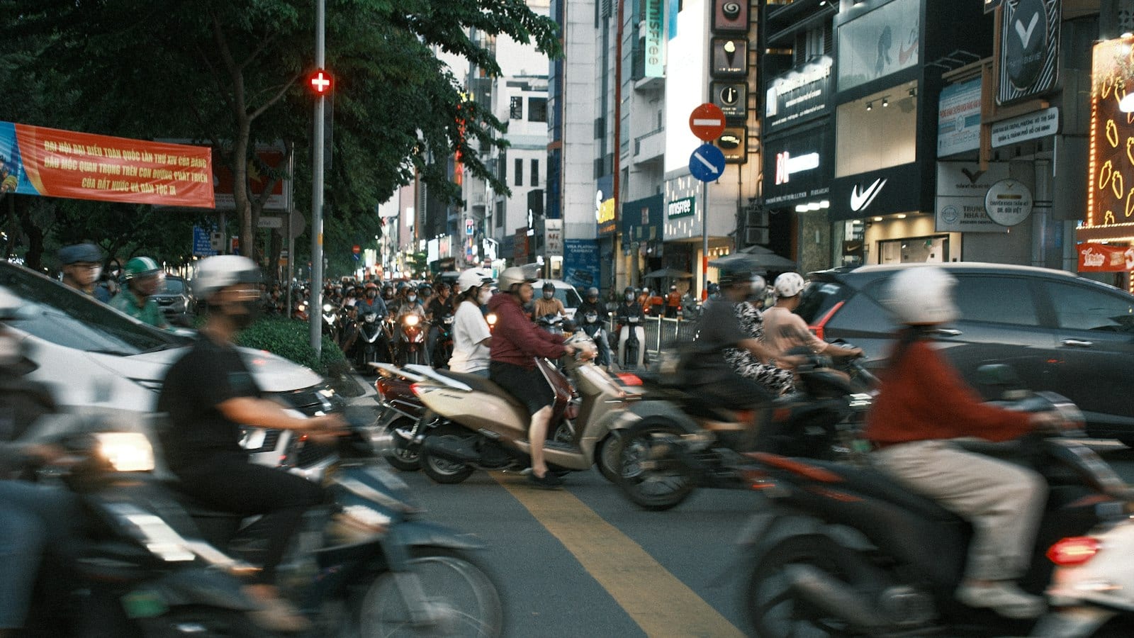 A city junction at dusk packed with riders, a red Party congress banner strung between trees on the left and shopfronts lit along the right