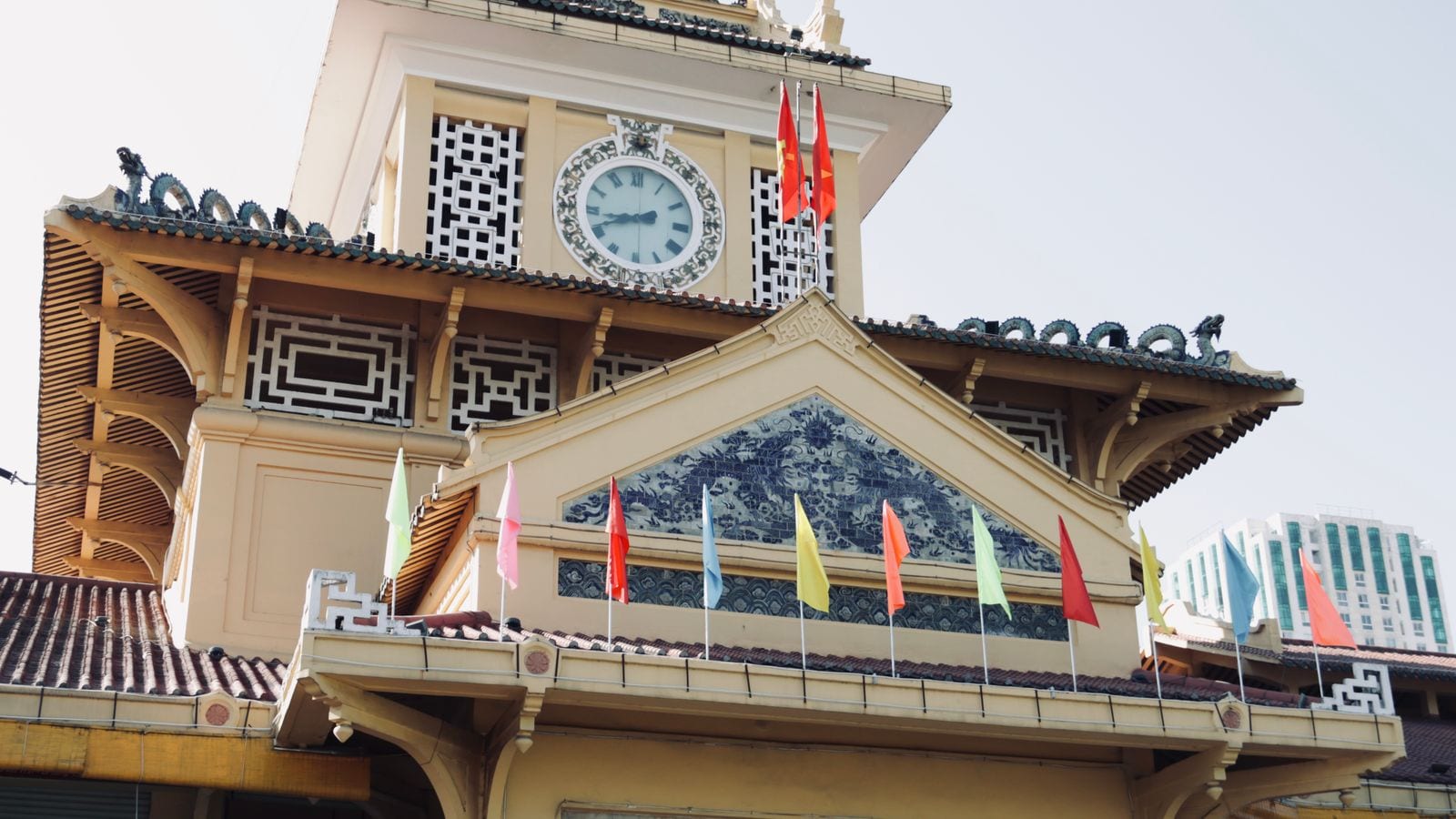 The clock tower of Binh Tay market in Cholon, tiled roof ridges ending in dragons, a blue and white ceramic panel in the gable and small flags along the parapet