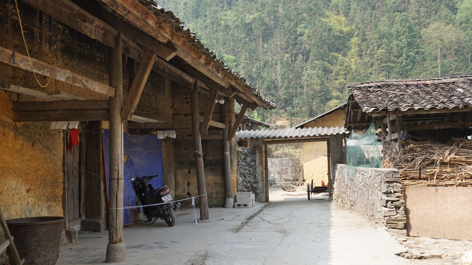 A lane between old rammed-earth houses in a highland village, tiled roofs on timber posts, a drystone wall opposite and maize stalks stacked against it