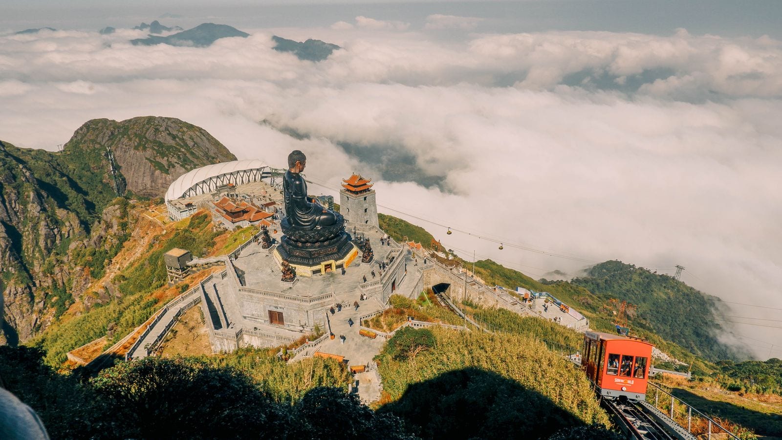 The summit complex on Fansipan seen from above the cloud line: a seated bronze Buddha on a terraced platform, a tiered tower beside it and the red funicular car climbing the last stretch
