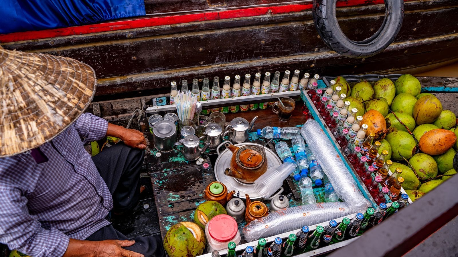 A vendor in a conical hat sitting in a boat loaded with young coconuts, rows of soft drinks and a stack of teapots, tied alongside a larger hull