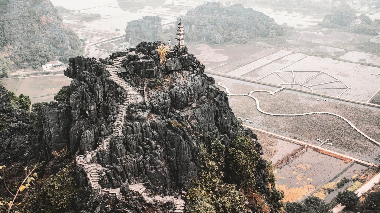 The stone stairway at Hang Mua climbing a black limestone outcrop to a small pagoda at the top, flooded fields and karst spreading out below