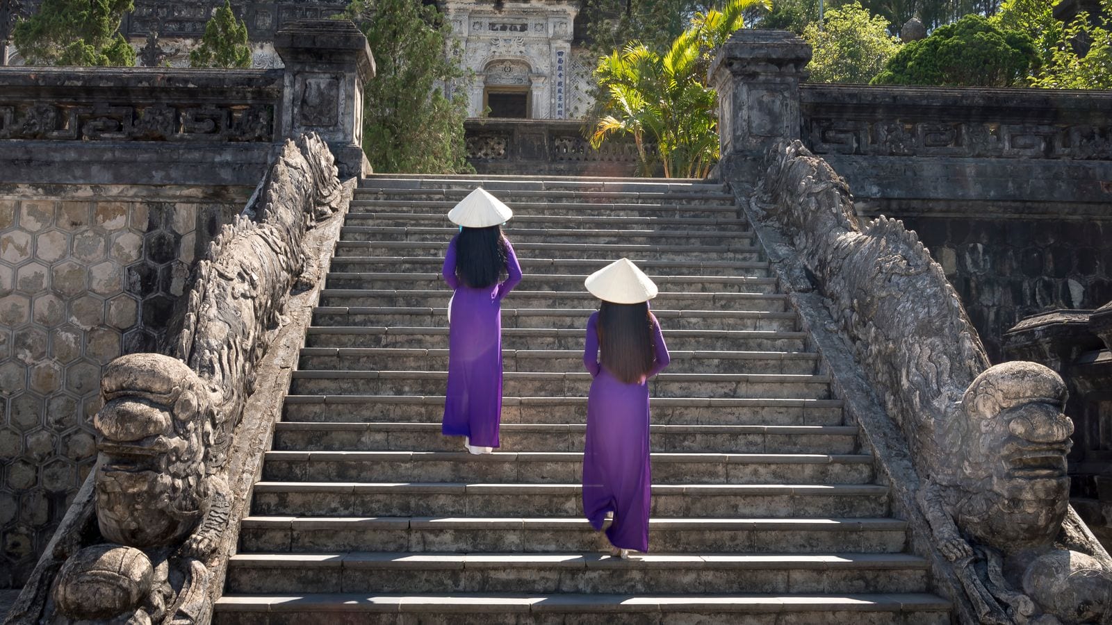 Two visitors in purple ao dai and conical hats climbing the stone stairway at the Khai Dinh tomb between carved dragon balustrades
