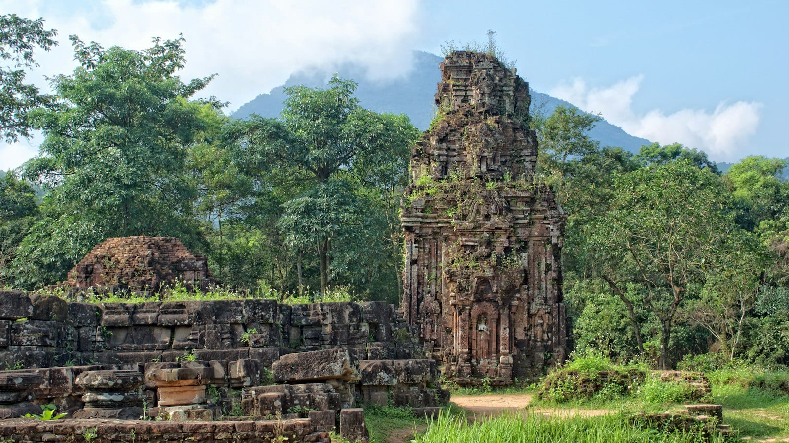 A Cham brick tower standing in cleared jungle with plants growing out of its top, a fallen wall of carved blocks in front of it and a forested mountain behind