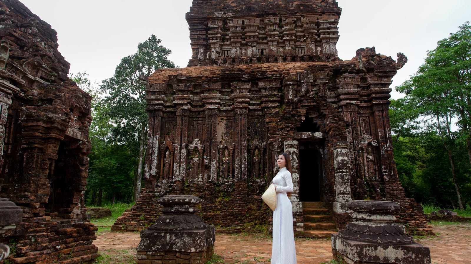 A Cham brick sanctuary standing among trees, its carved pilasters and tiered roof eaten into by weather, a visitor in a white ao dai at the doorway for scale