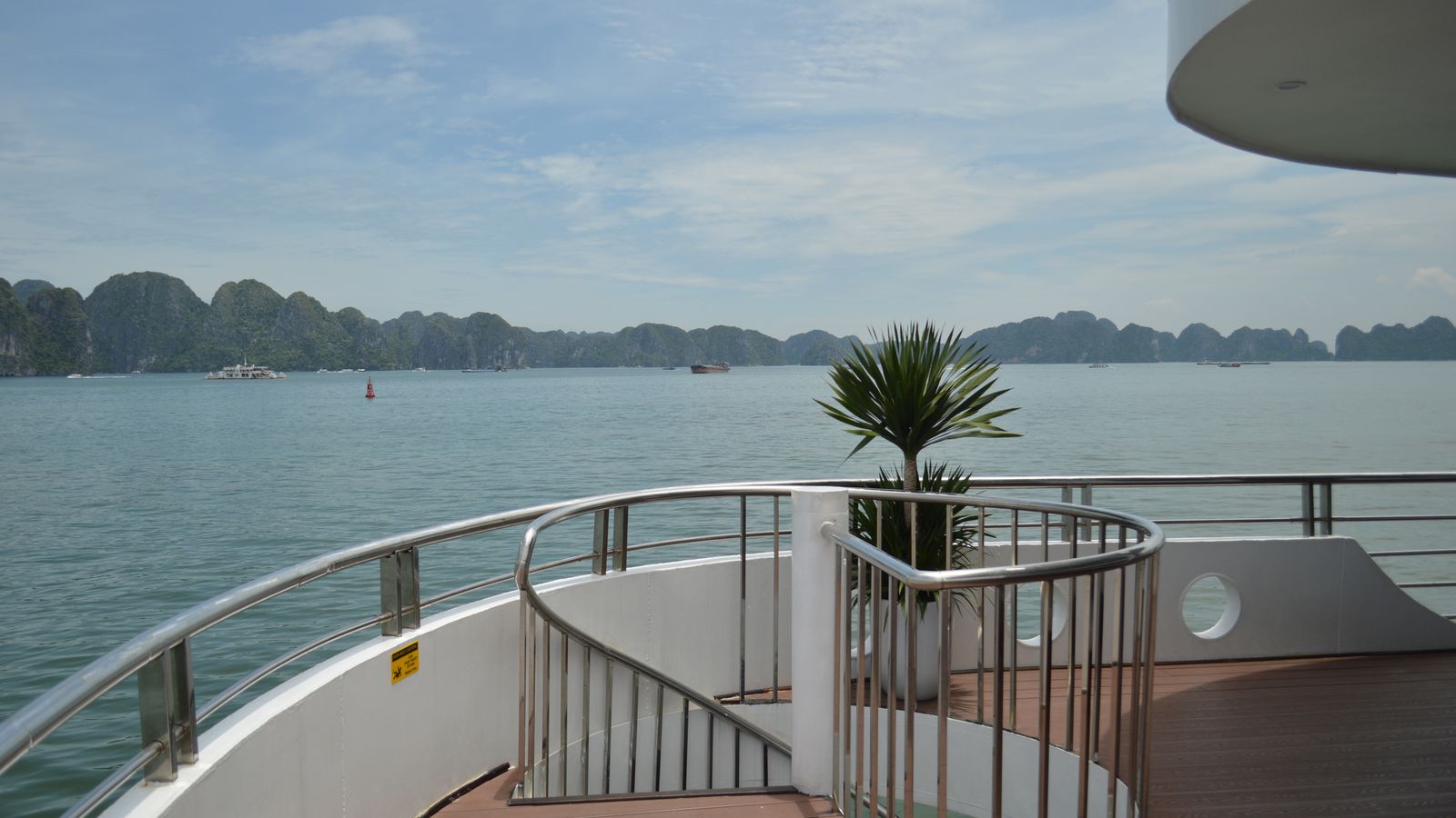 The empty sun deck of a cruise boat, stainless railings and a potted palm, with limestone islands strung across the bay beyond