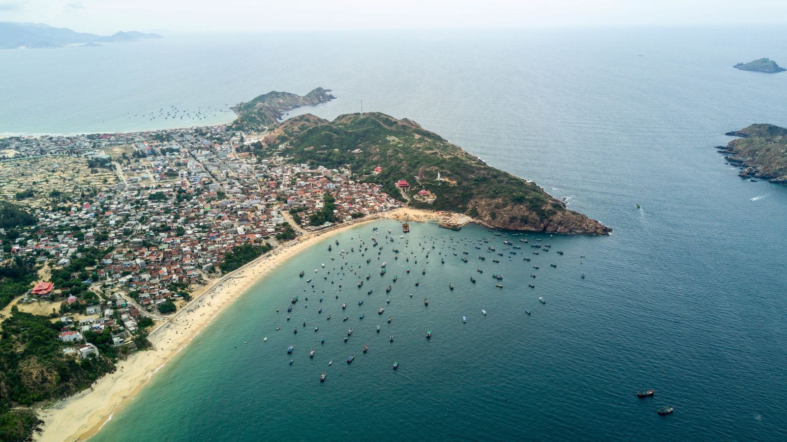 A fishing town packed onto a headland from the air, tiled roofs running down to a curve of sand and a hundred boats moored off the beach