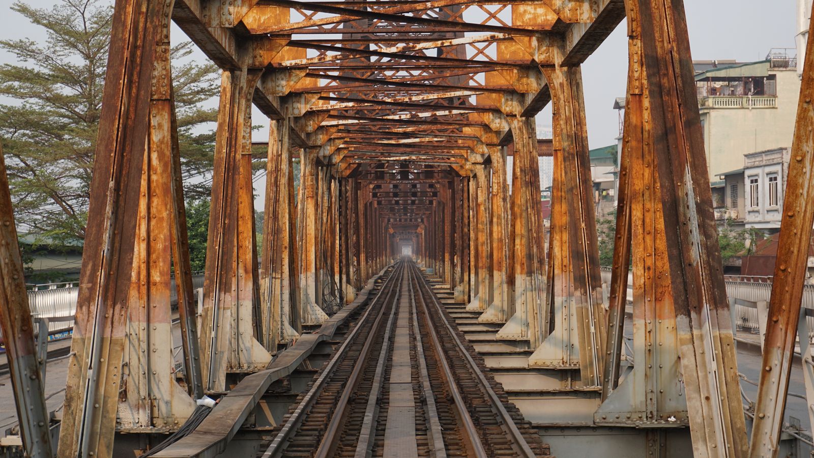 The deck of an old steel railway bridge, rusted lattice girders receding to a point over a single track, with shophouse balconies close on the right