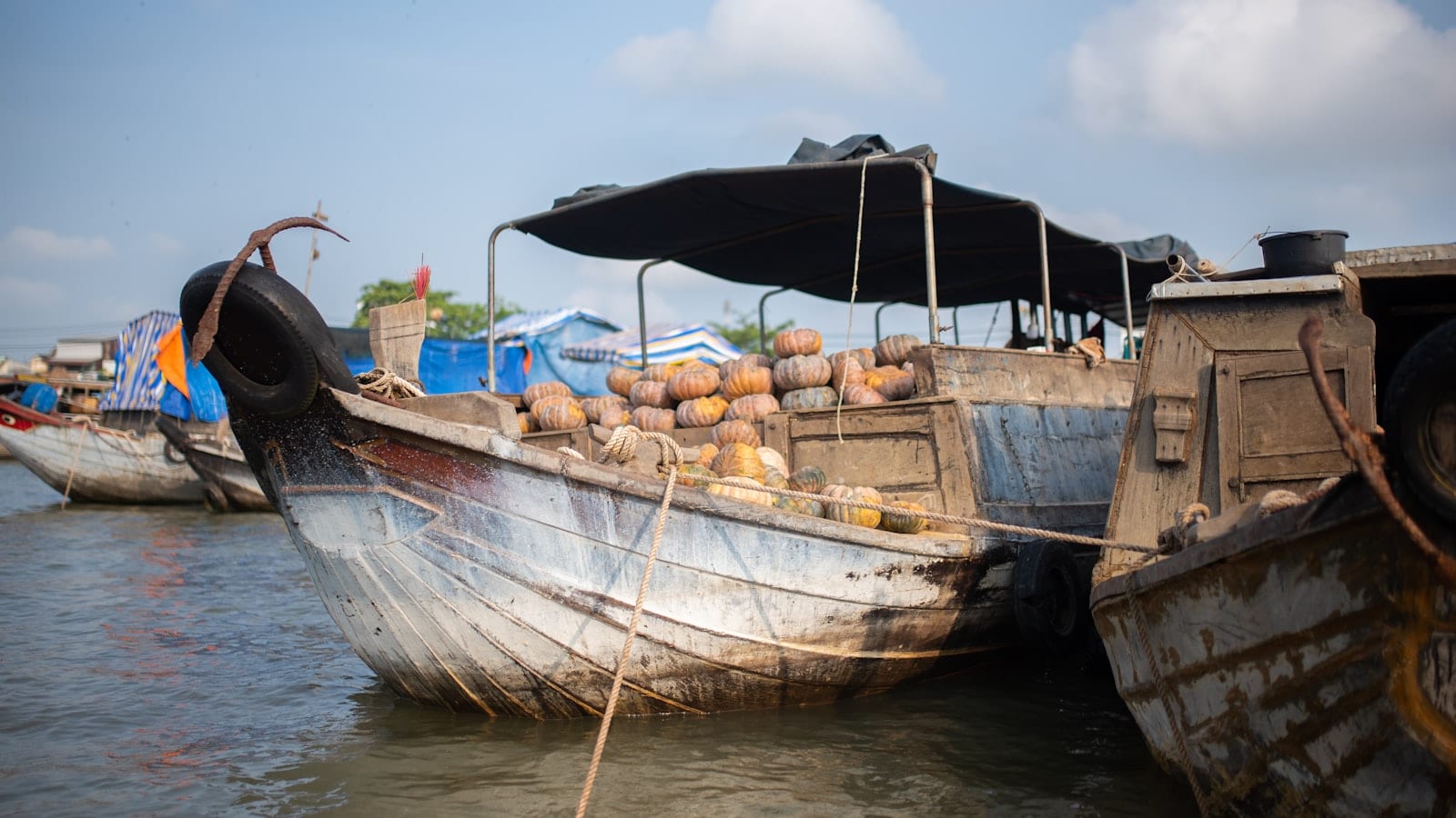 A wooden cargo boat piled with pumpkins under a black canopy, moored hull to hull with another boat on a brown river