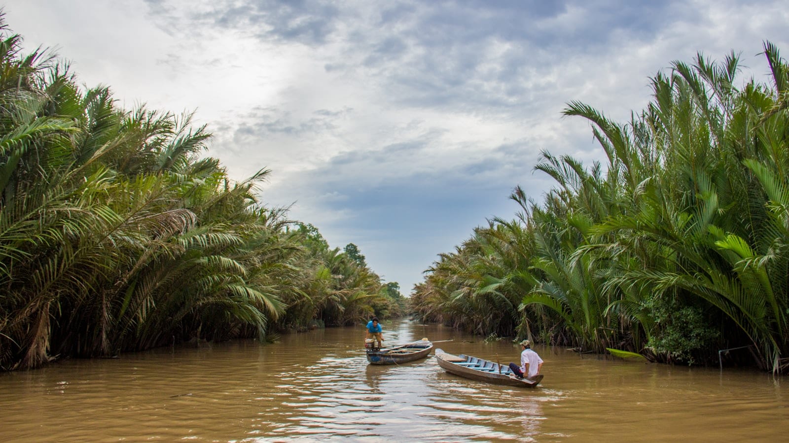 Two wooden boats on a narrow brown channel lined with dense water palms