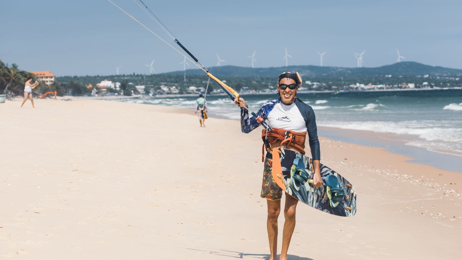 A kitesurfer walking up the sand at Mui Ne with his board, wind turbines standing along the ridge behind the bay