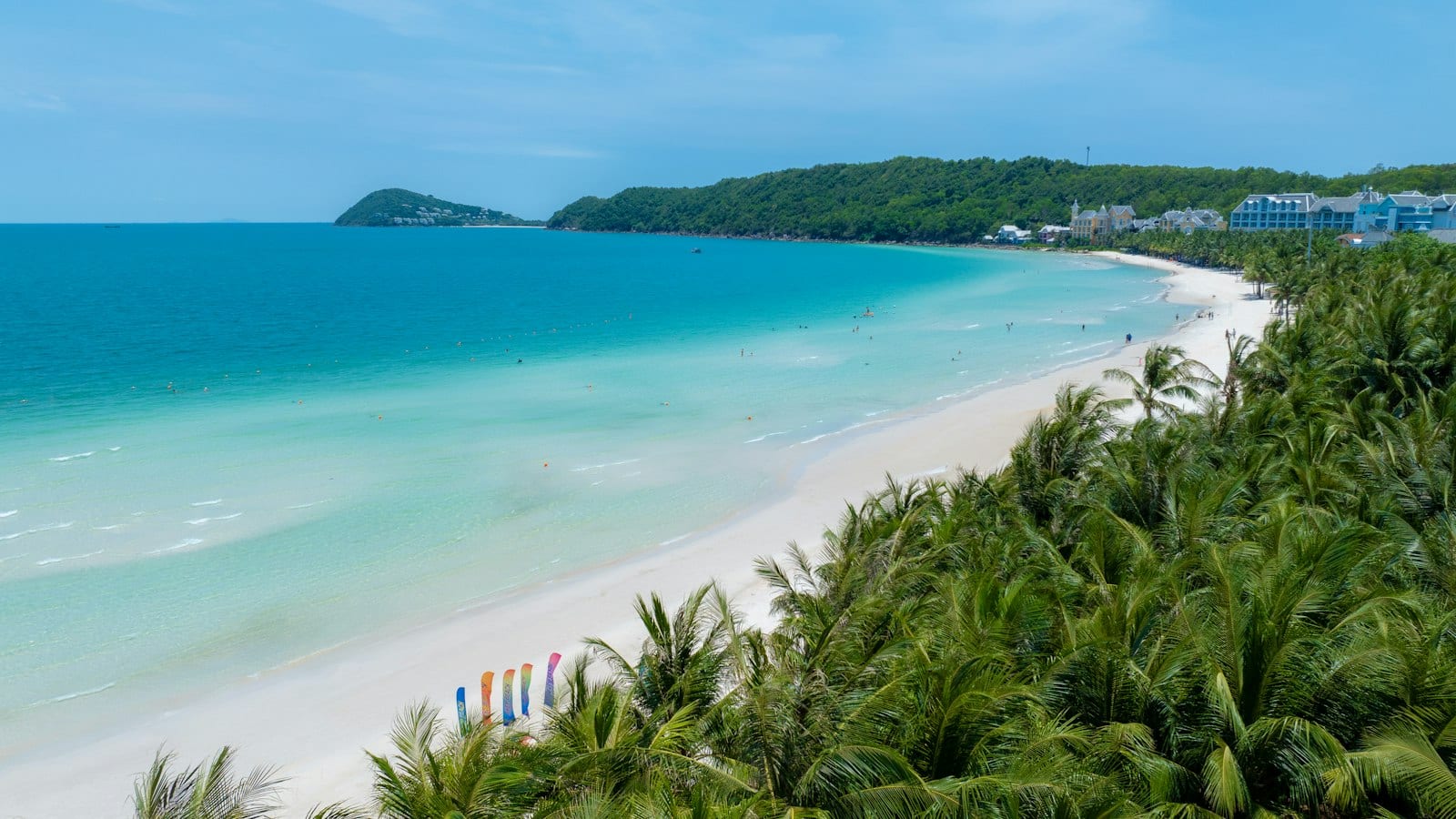 White sand curving along shallow turquoise water on southern Phu Quoc, palms in the foreground and the pastel blocks of a resort along the headland