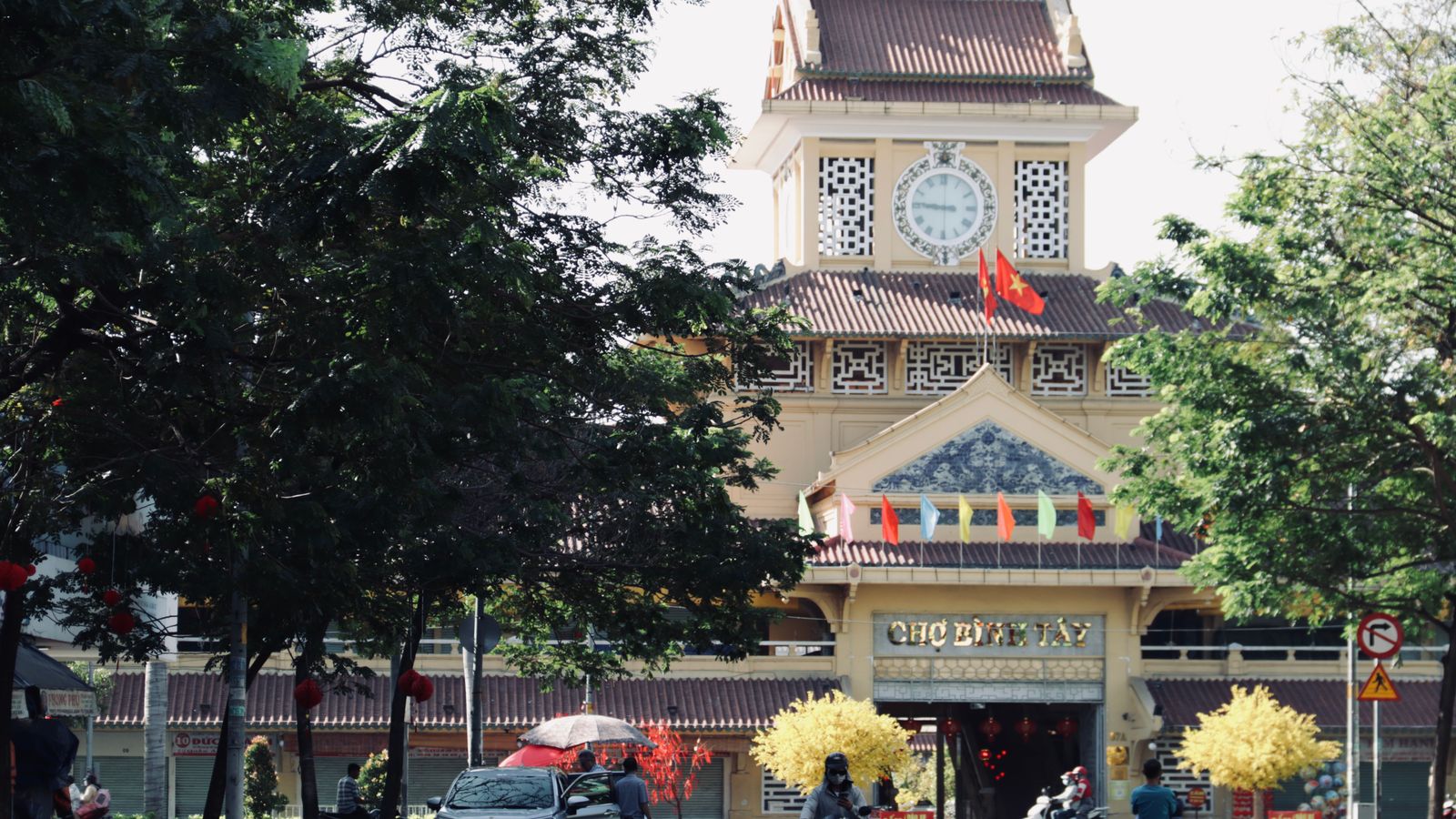 The yellow clock tower of Binh Tay market seen through street trees, CHO BINH TAY over the entrance, red flags on the roof and yellow apricot blossom set out for Tet below