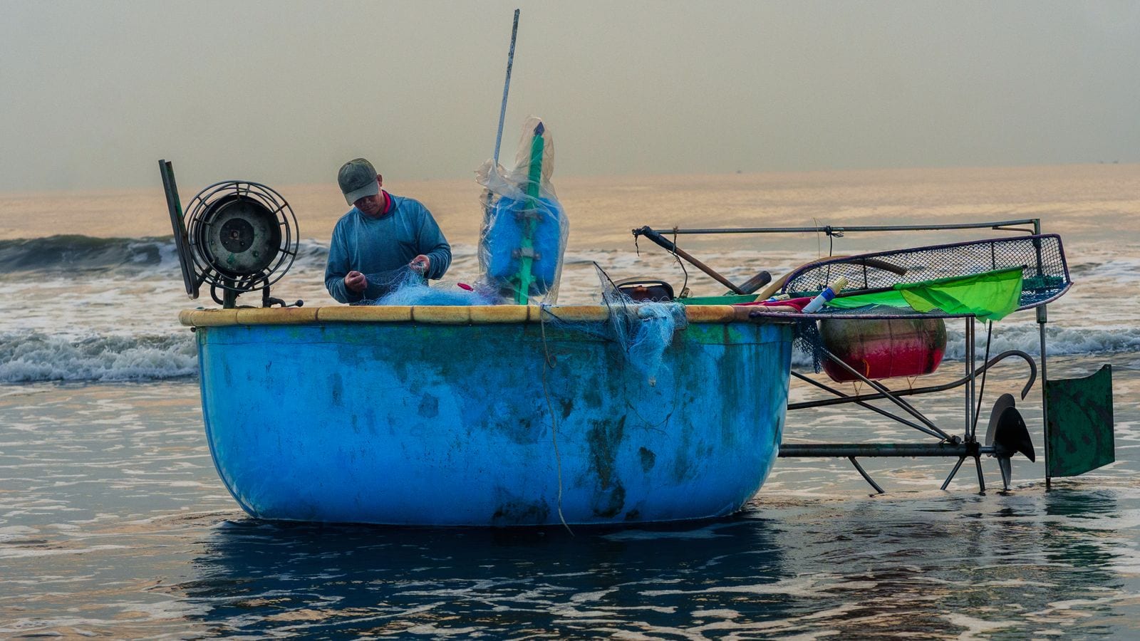 A man sorting net inside a blue round basket boat in the shallows, an outboard on a long frame clamped over the side