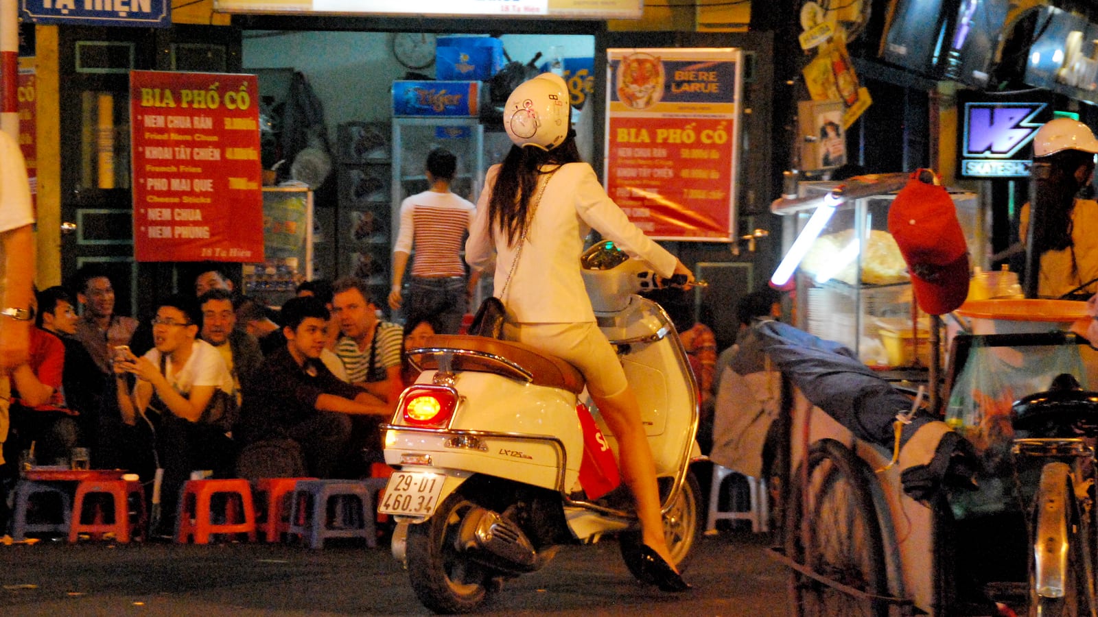 The corner of Ta Hien at night: drinkers on red and blue plastic stools under beer signs, a scooter riding past and a food cart lit up on the far pavement