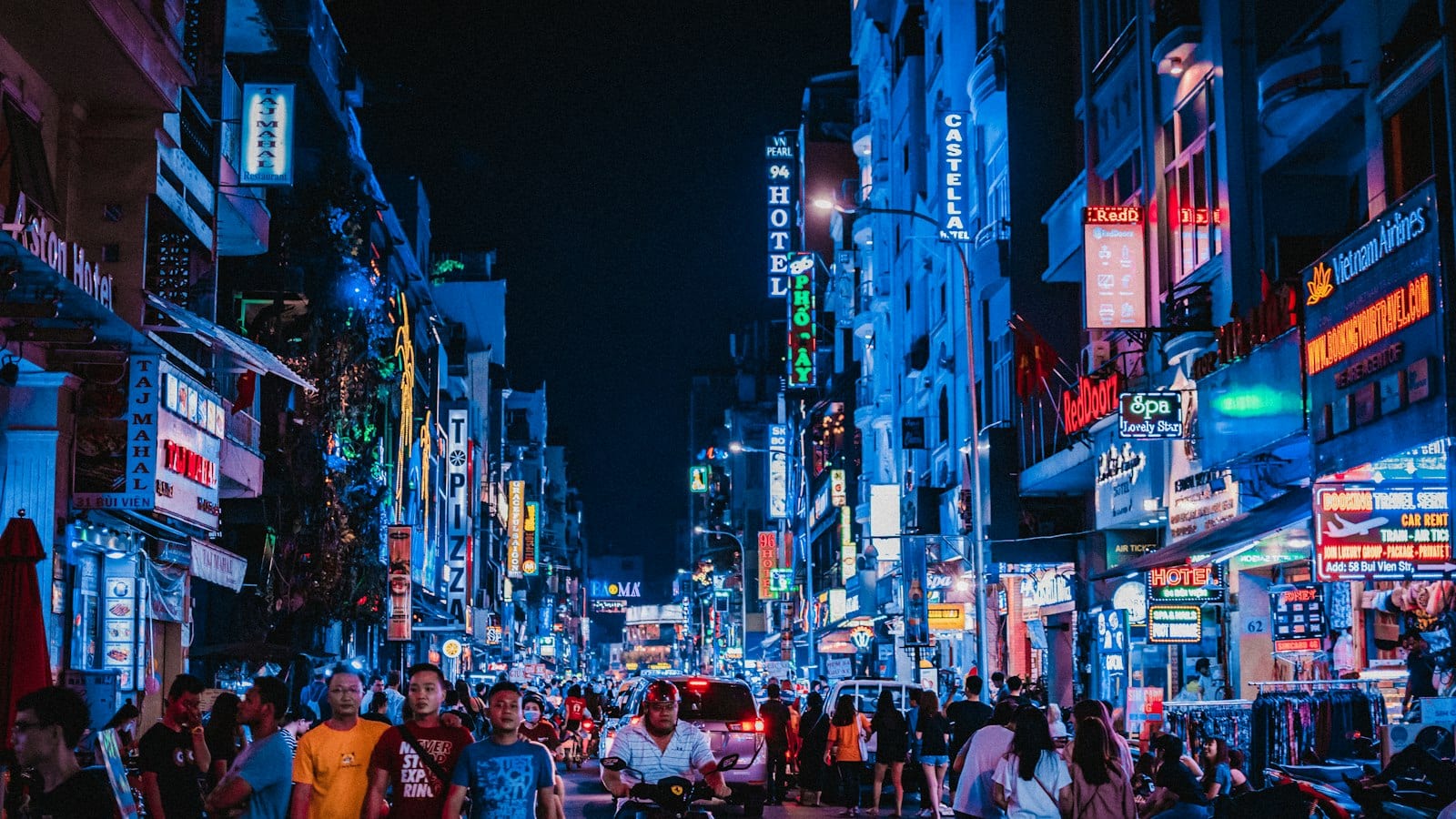 A backpacker street at night under two walls of stacked neon — hotels, spas, pizza, air tickets — with the crowd spilling off the pavements into the road
