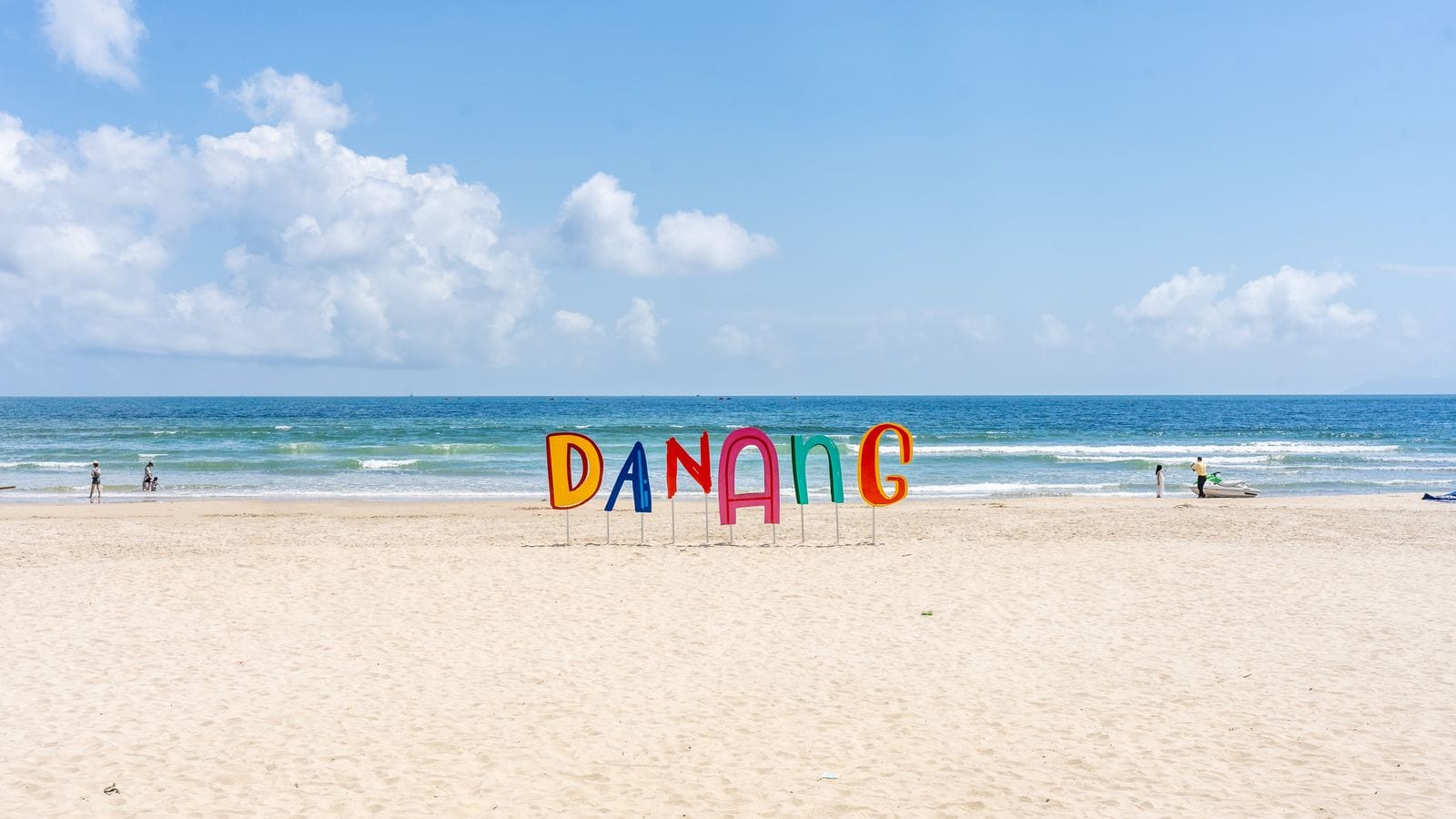 A wide empty beach with the word DANANG standing in coloured letters on the sand, two people at the waterline and nobody else in the frame