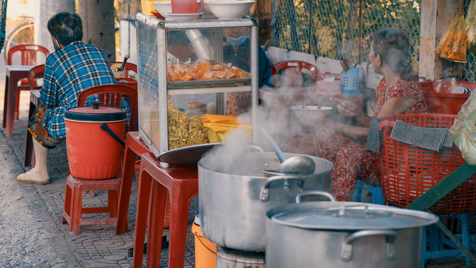 A pavement noodle stall at first light: two steel pots steaming on the kerb, a glass case of noodles and herbs, red plastic stools, and the seller passing a bowl across