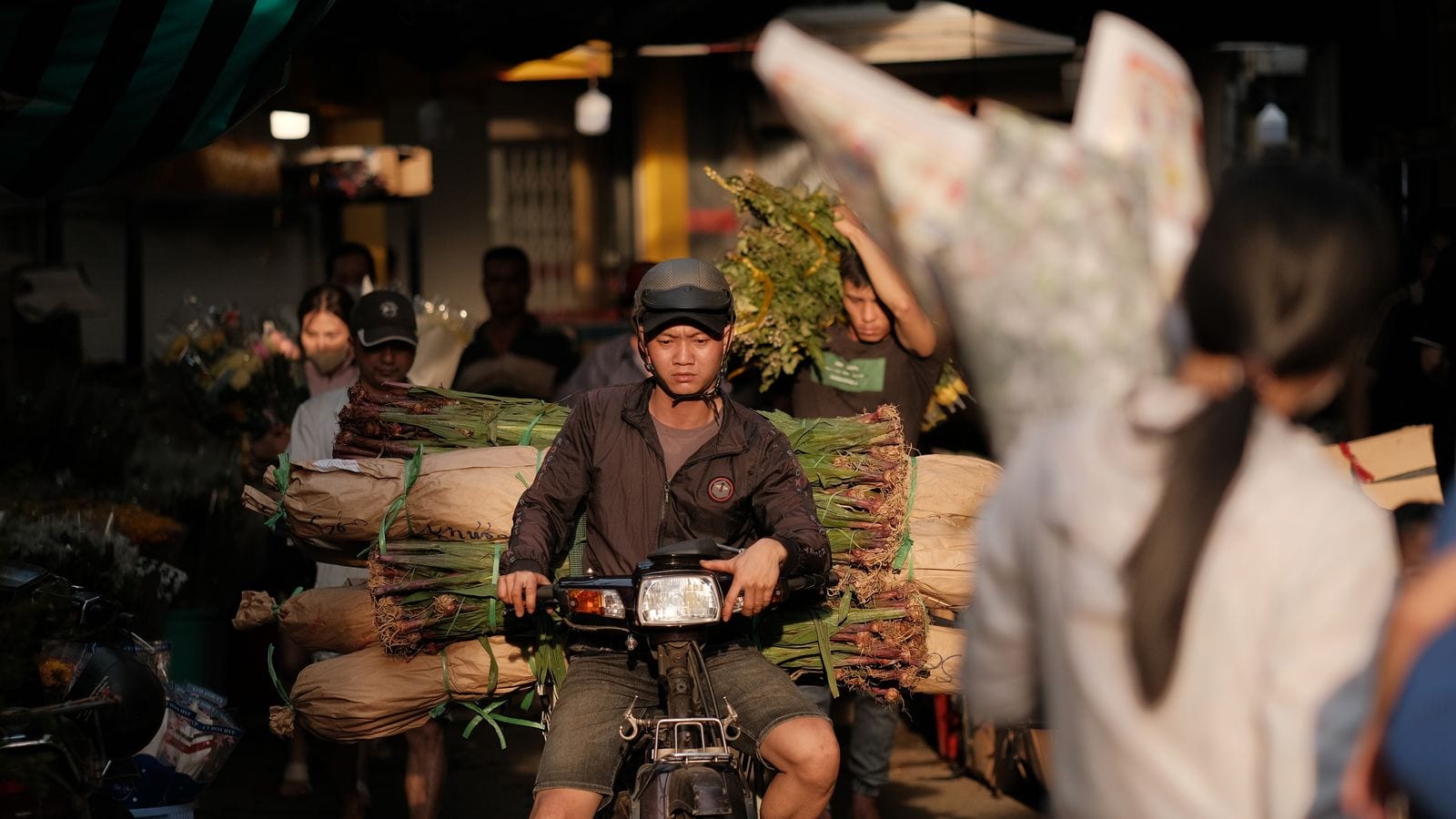 A rider carrying two long paper-wrapped bundles of flowers sideways across the back of his motorbike through a covered market, with porters walking bundles out behind him