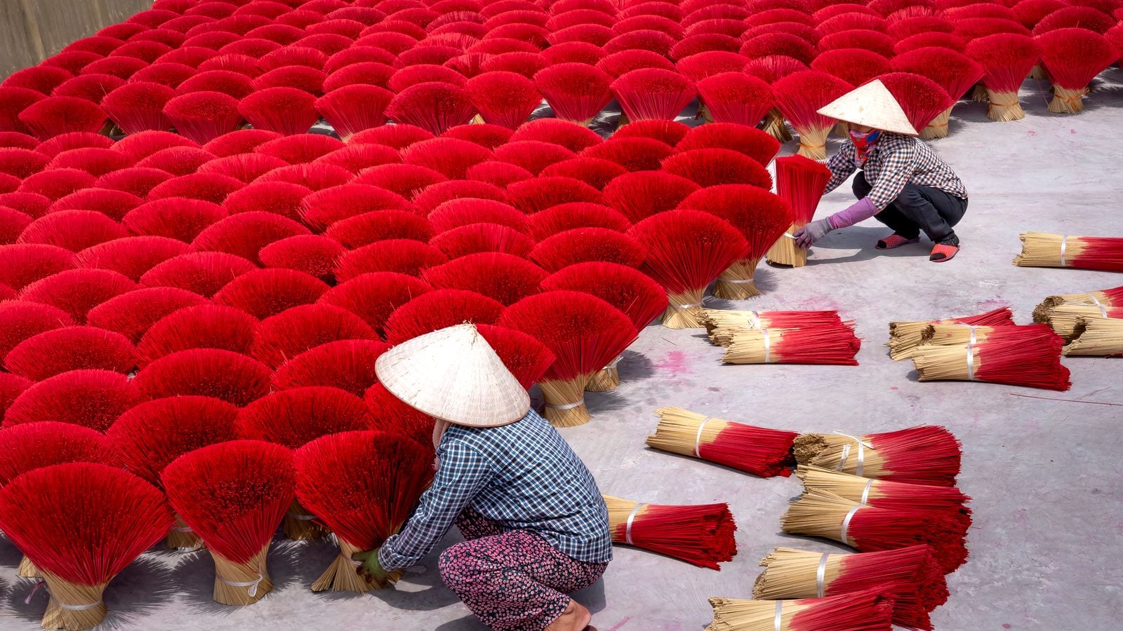 Two women in conical hats setting out bundles of incense sticks to dry, the dyed red ends fanned open across the whole yard