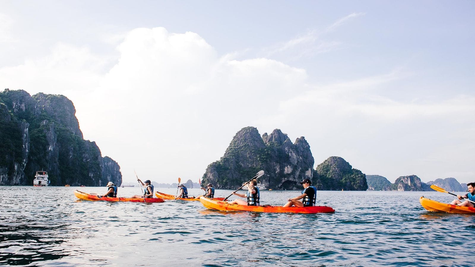 Half a dozen orange kayaks on flat water between limestone islands, with a white cruise boat anchored in the channel behind them