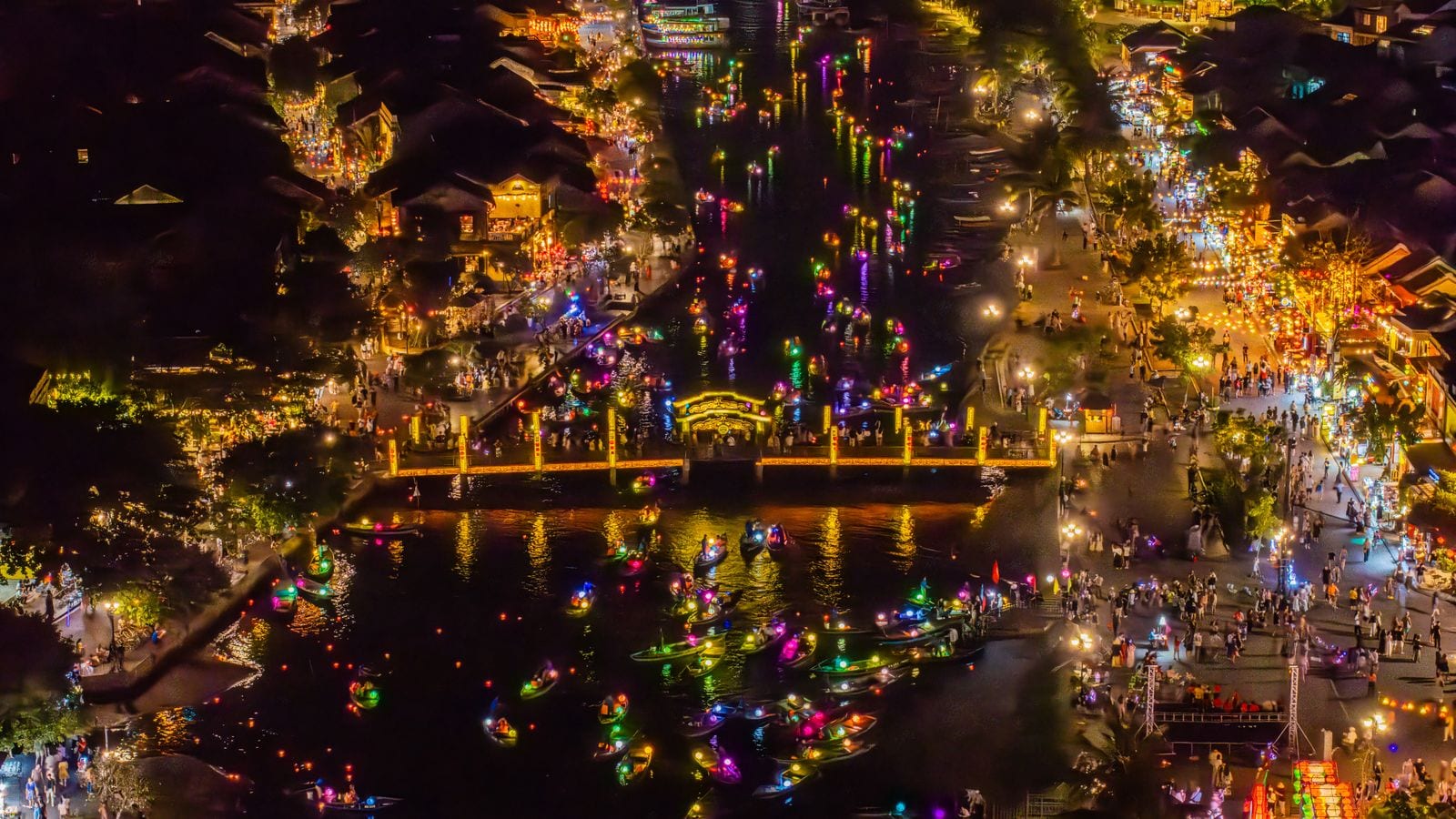 Hoi An from above on a lantern night: coloured lanterns strung over the old town, boats carrying paper lanterns down the river and the footbridge lit end to end