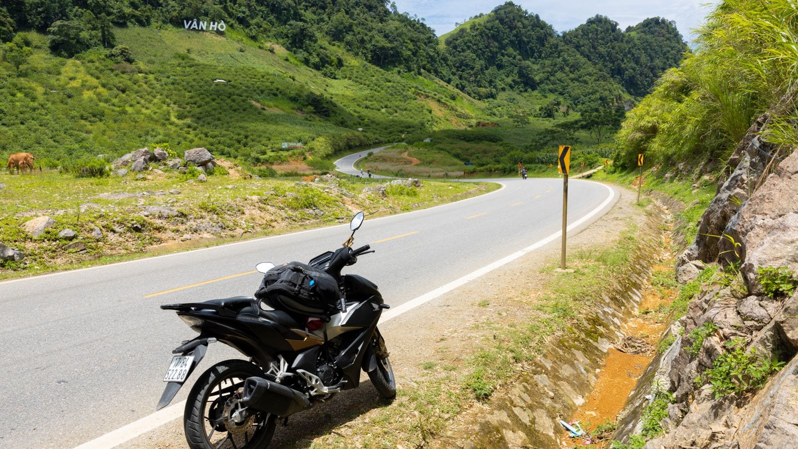 A motorbike with a rucksack strapped to the seat parked on the verge of a mountain road, the tarmac curving down through green hills with VAN HO spelled out on the hillside opposite