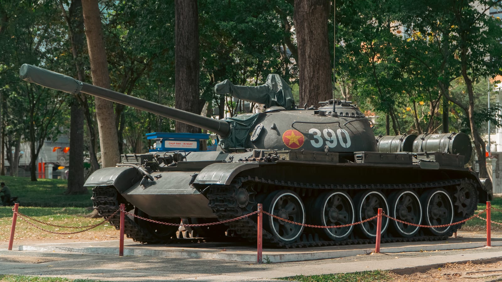 A T-54 tank painted with the number 390 and a gold star standing chained off under trees, a photo-printing stall set up behind it