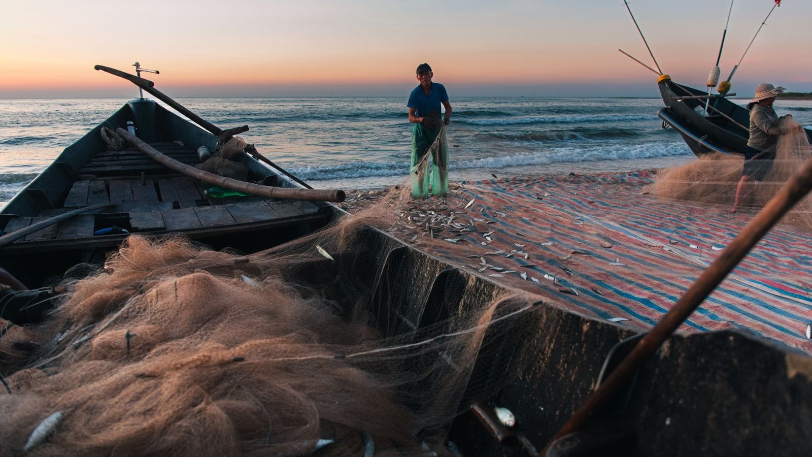 Fishermen shaking small fish out of a net on the sand at dawn, one standing knee-deep in the surf beside a wooden boat, nets heaped across the whole foreground