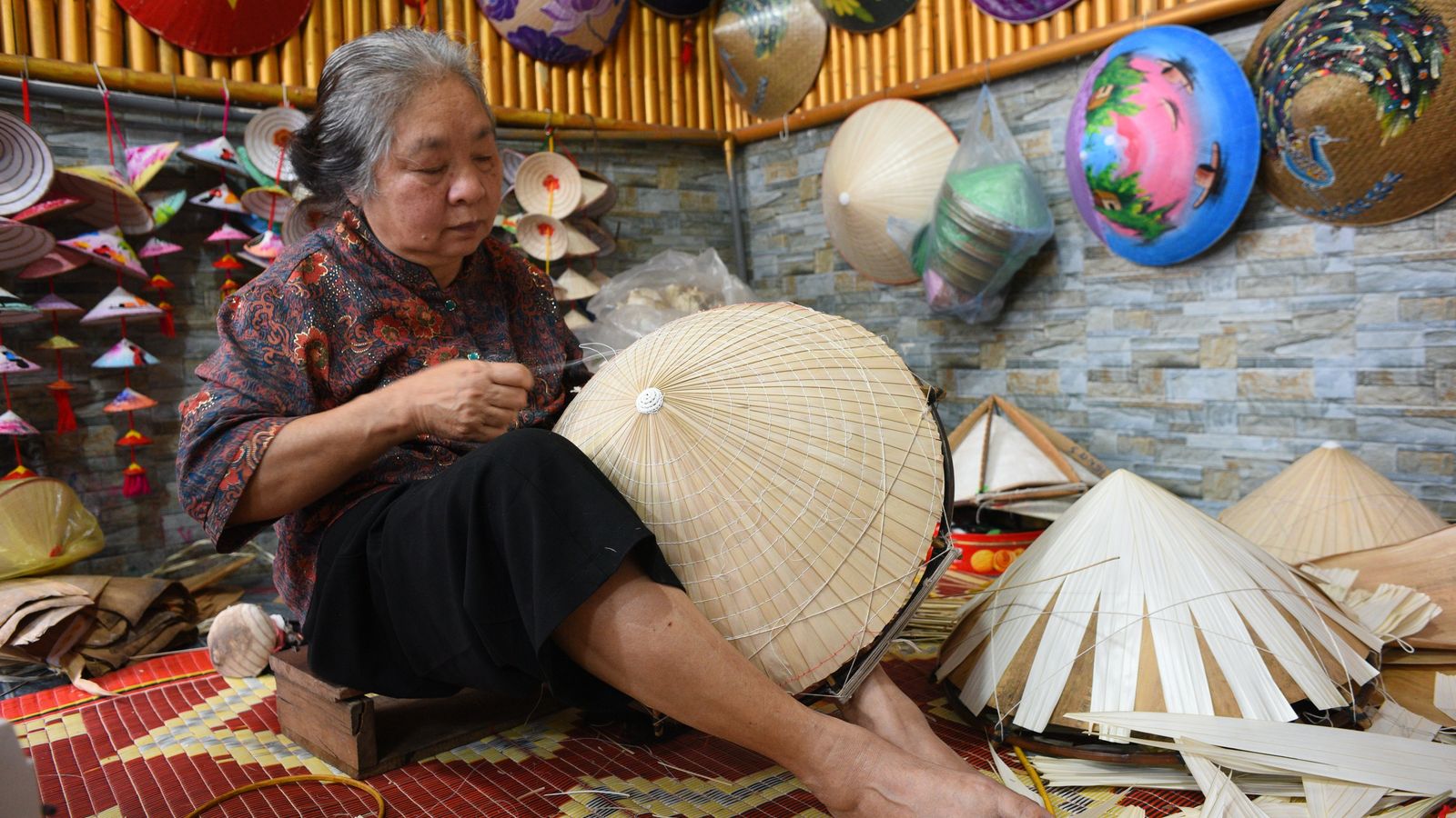 A woman stitching palm leaf onto the bamboo frame of a conical hat, finished hats hanging on the wall behind her and cut leaf stacked on the mat