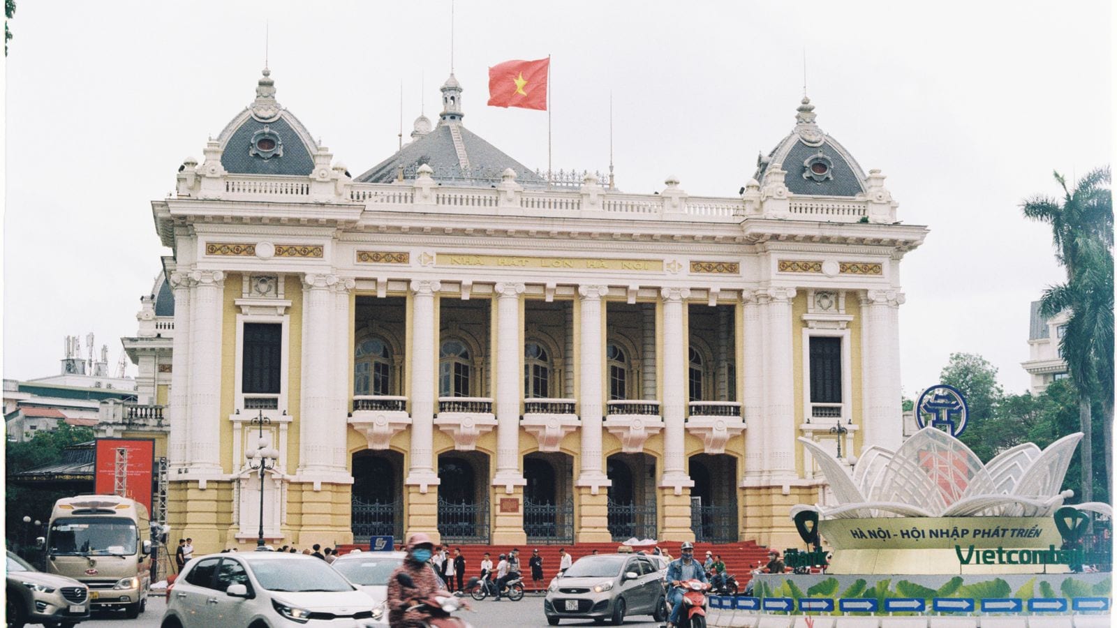 The Hanoi Opera House across a busy junction, NHA HAT LON HA NOI lettered above the colonnade, a national flag on the roof and traffic crossing in front