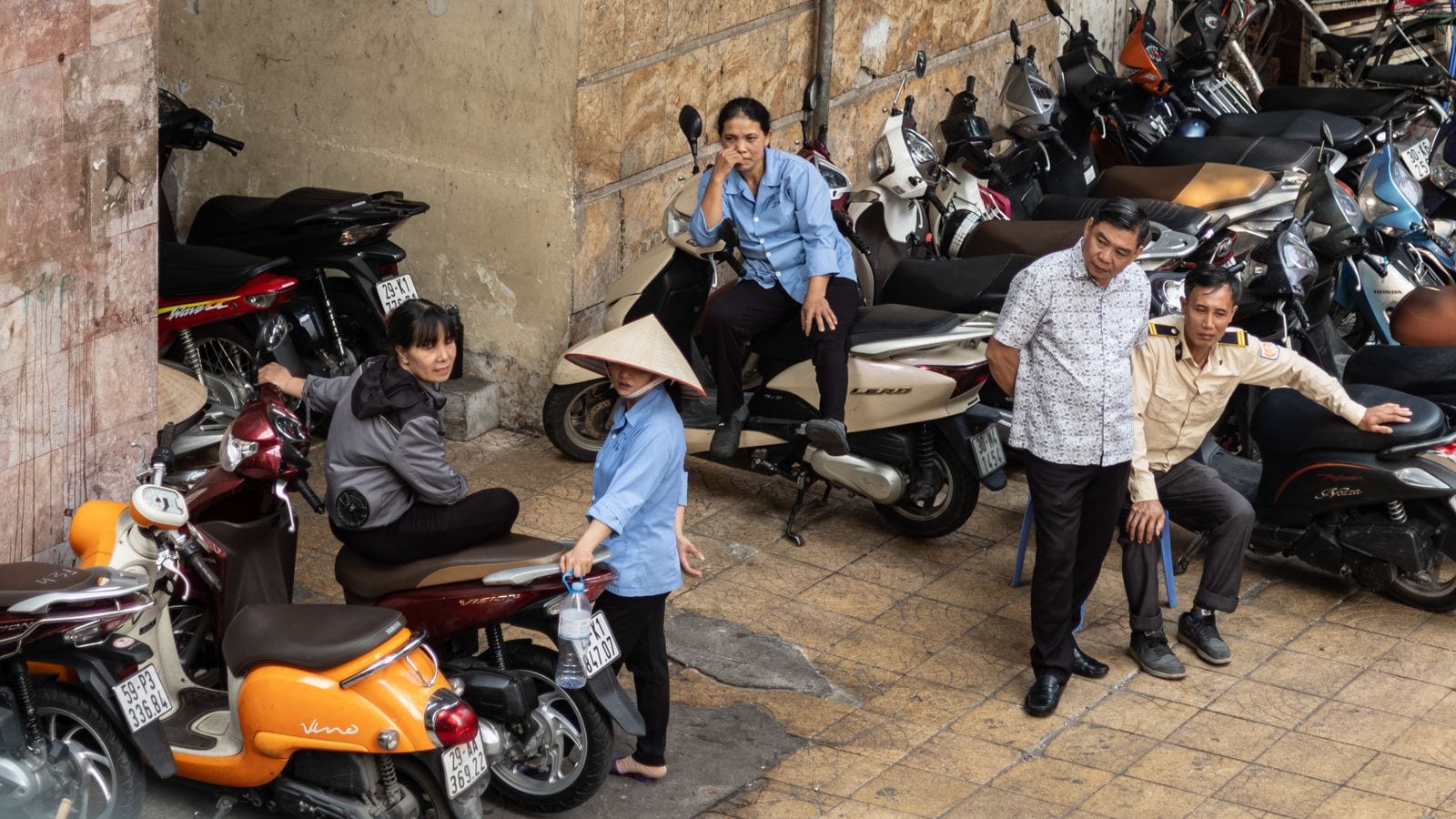 A pavement packed end to end with parked scooters, the attendants sitting on the saddles with nothing left to walk on but a metre of tiles