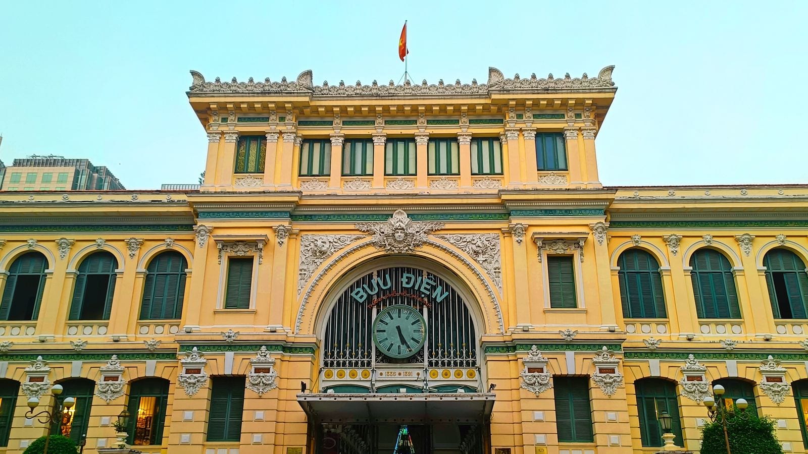 The front of the Saigon central post office: ochre plaster, green shutters, a clock under the arch and BUU DIEN THANH PHO HO CHI MINH in metal letters above it