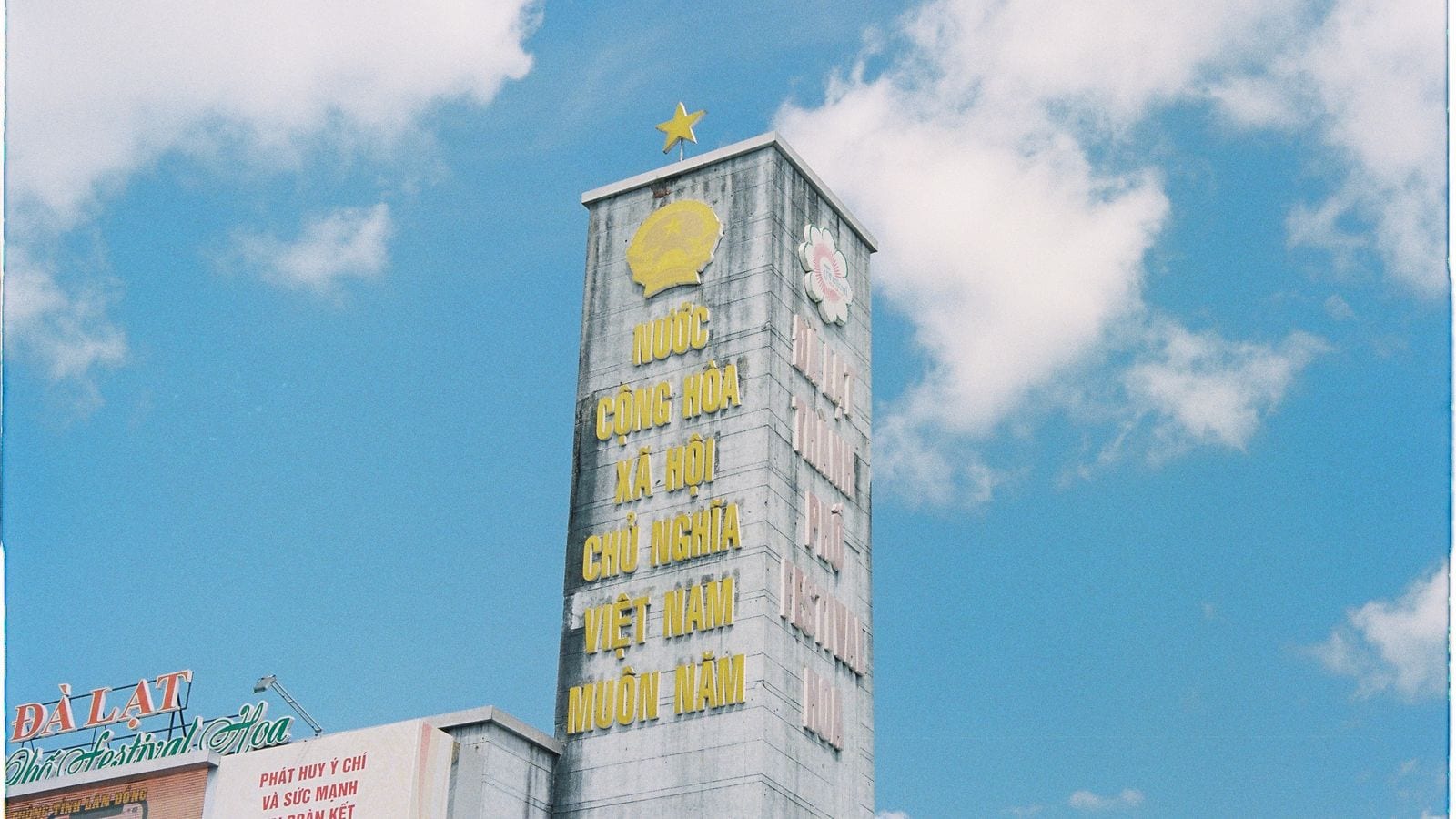 A concrete tower with a gold star on top and the state emblem below it, the slogan NUOC CONG HOA XA HOI CHU NGHIA VIET NAM MUON NAM running down the face in yellow letters