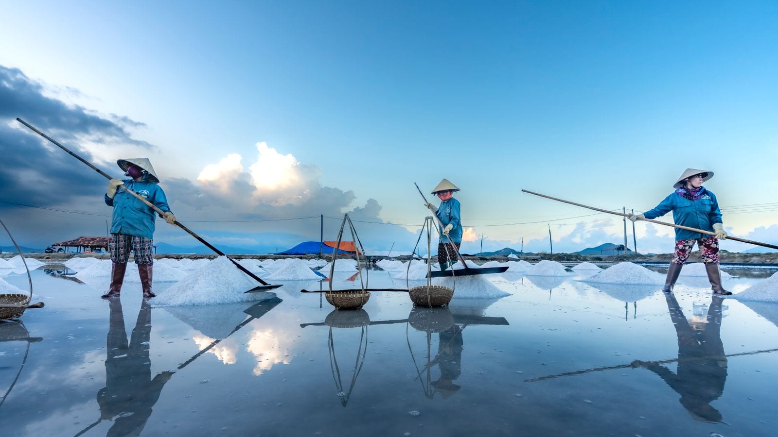 Three workers in conical hats raking salt into cones on a flooded pan, with carrying baskets and a shoulder pole standing between them