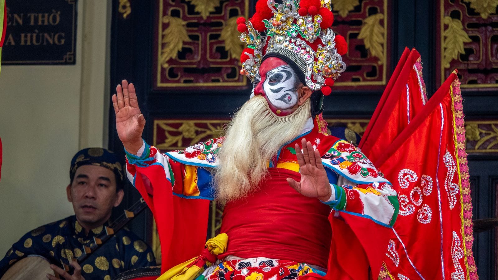 A tuong performer in a red robe and jewelled headdress, face painted red and white with a long white beard, a musician with a two-string lute seated behind him