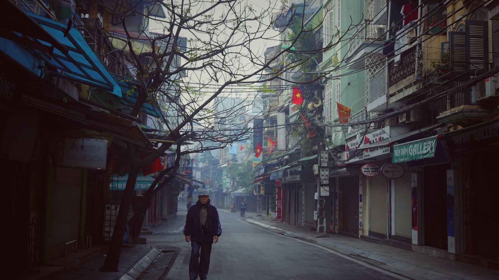 A narrow street of tube houses early in the morning, shutters still down, national flags hung out over the shopfronts and cables strung across between them