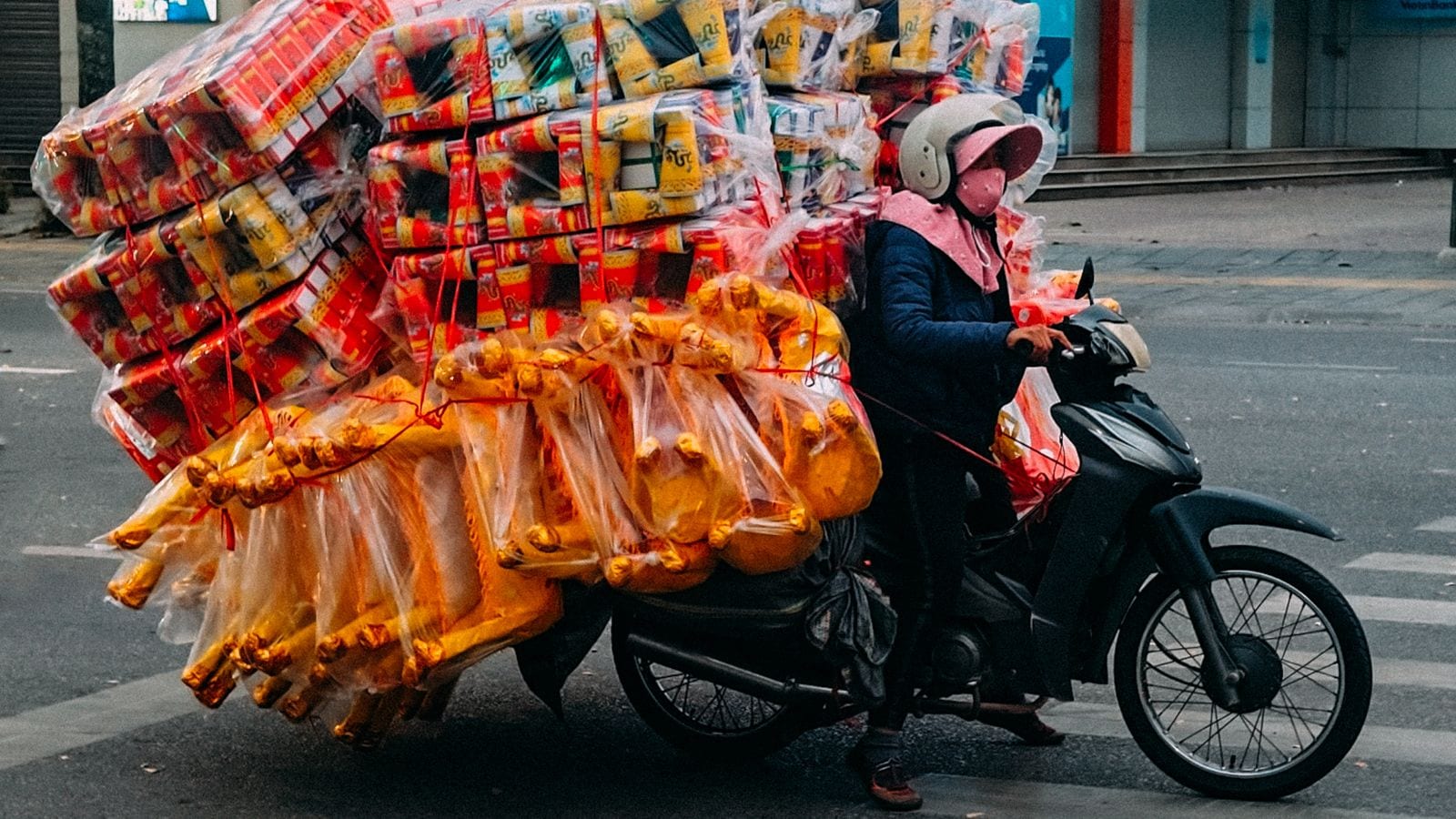 A rider stopped at a junction with votive paper goods stacked head-high behind the saddle — bundles of gold ingots and boxed offerings in clear plastic, tied down with red twine