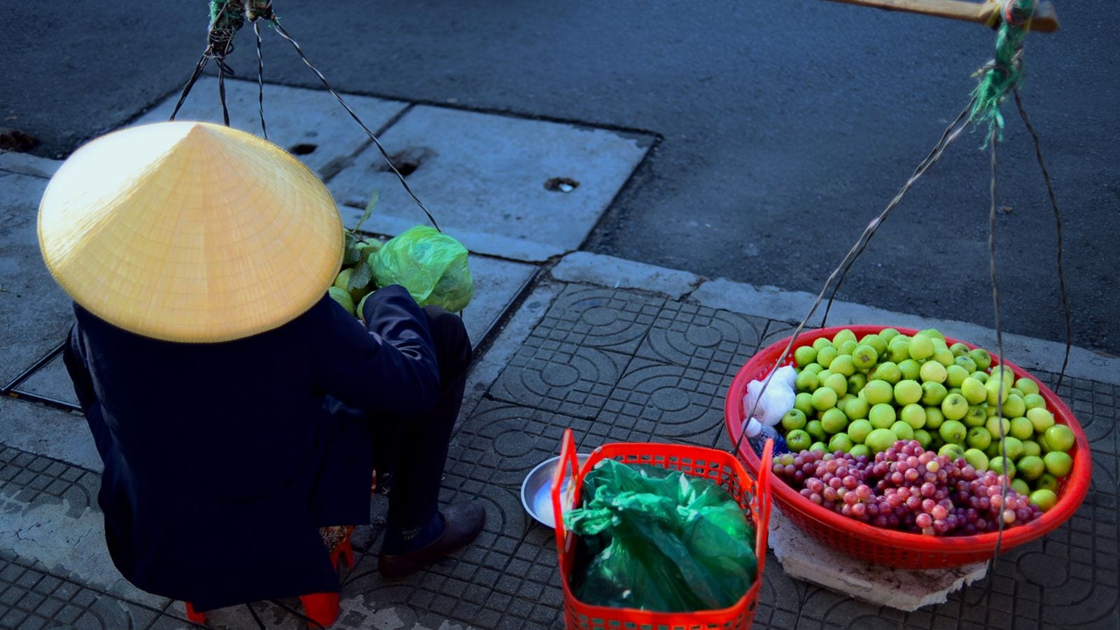 A vendor in a conical hat sitting on the kerb between the two baskets of her shoulder pole, one full of green apples and grapes