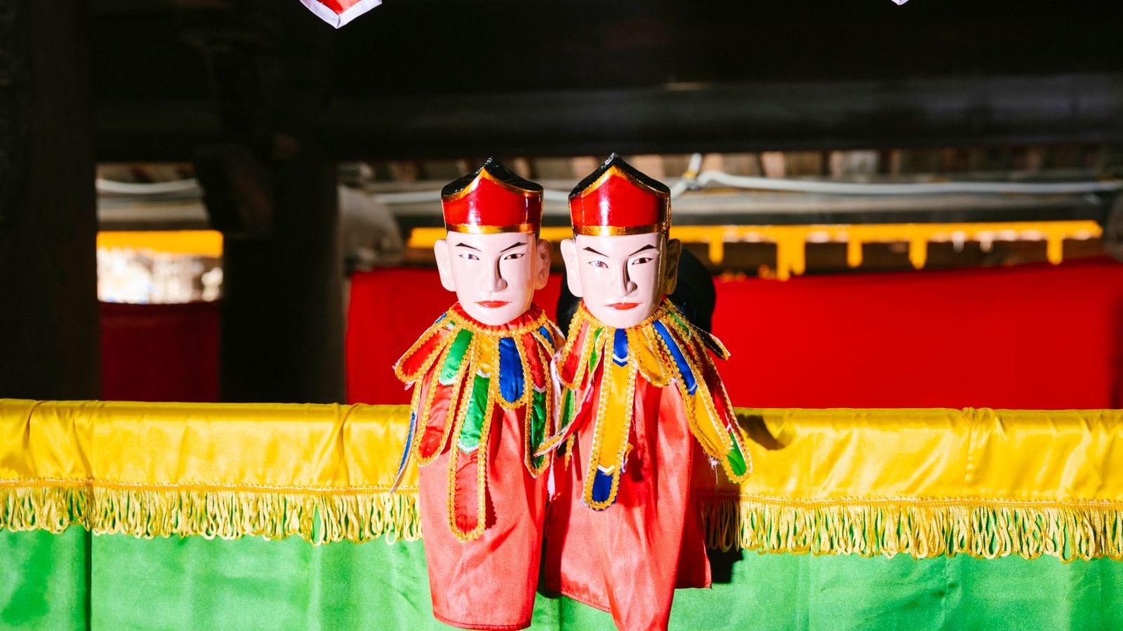 Two lacquered water puppets in red robes and gold-trimmed collars standing above the water in front of the green and yellow curtain they come out from