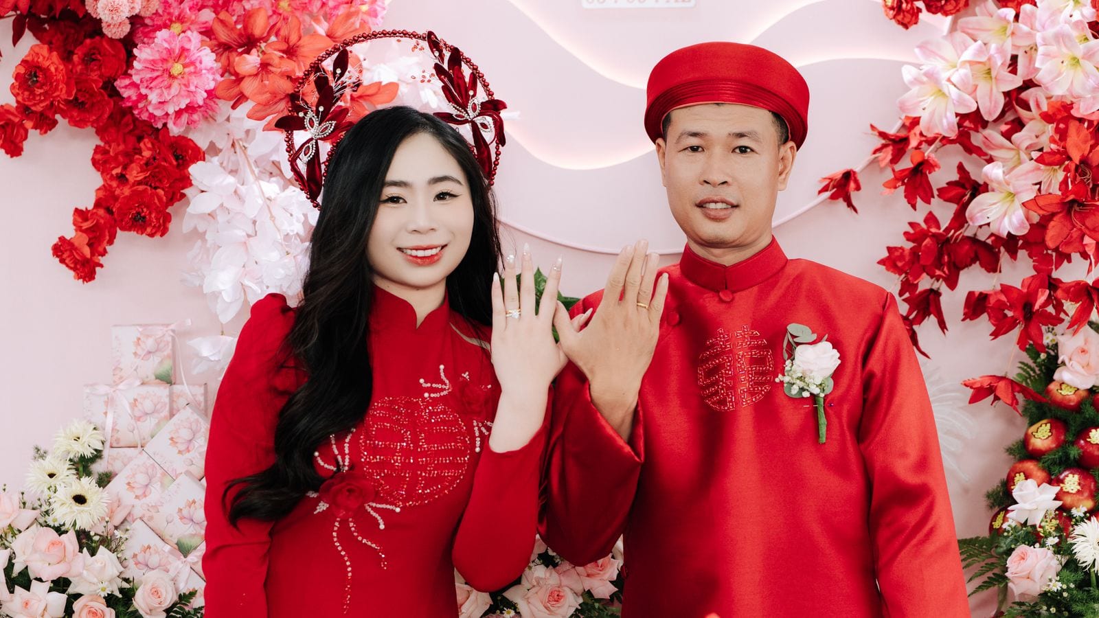 A bride and groom in matching red ao dai in front of a flower wall, she in a beaded headdress, he in a red khan dong, both showing their rings