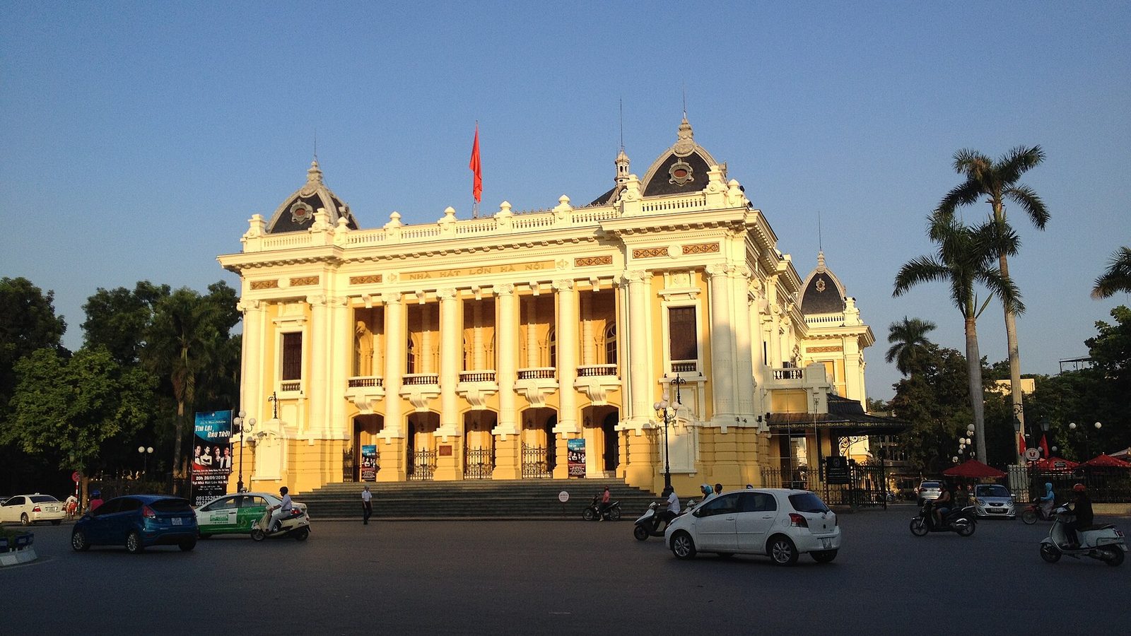 The yellow and white facade of the Hanoi Opera House in late afternoon sun, NHA HAT LON HA NOI above the columns, palms on the right and scooters crossing the square